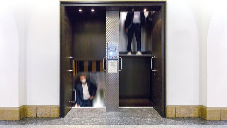 Mann, der in einem Paternoster auf und ab fährt. Ein Paternoster ist ein Aufzug mit zwei nebeneinander liegenden Schächten, die sich permanent auf und ab bewegen.